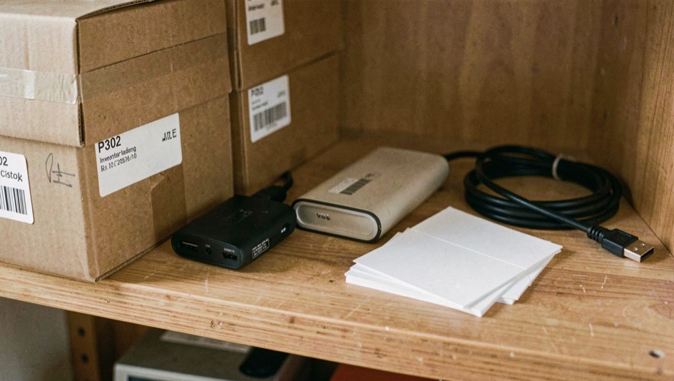 Close documentary photograph of storage boxes, a compact external drive, a cable and blank inventory cards on a wooden shelf, no legible writing, warm daylight. Distinct supporting card P302 2 composition for this editorial entry.