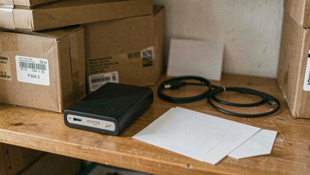 Close documentary photograph of storage boxes, a compact external drive, a cable and blank inventory cards on a wooden shelf, no legible writing, warm daylight. Distinct supporting card P304 2 composition for this editorial entry.