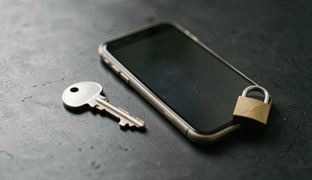 Documentary photograph of a metal key, a phone with a blank dark screen and a small padlock on a charcoal surface, no visible writing, controlled side light. Distinct lead composition for this editorial entry.