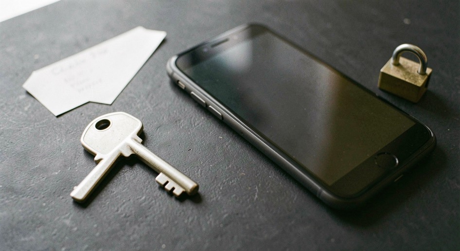 Documentary photograph of a metal key, a phone with a blank dark screen and a small padlock on a charcoal surface, no visible writing, controlled side light. Distinct supporting card P201 1 composition for this editorial entry.