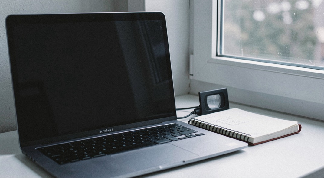 Documentary photograph of a laptop with a completely blank dark screen beside a notebook, a small privacy shutter and a window reflection, no readable interface, cool morning light. Distinct section Sicherheit 1 composition for this editorial entry.