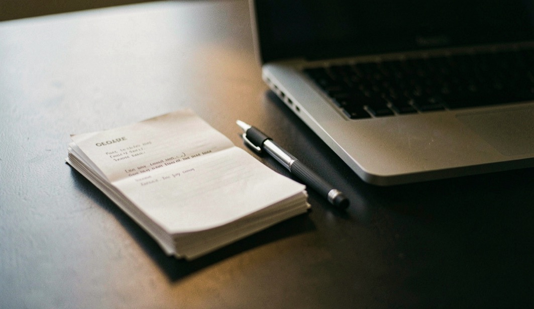 Documentary photograph of a small stack of paper drafts, a mechanical pencil and a compact laptop on a dark desk, no readable words, a single warm pool of light. Distinct lead composition for this editorial entry.