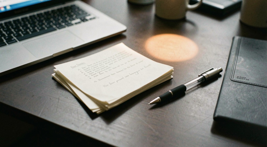 Documentary photograph of a small stack of paper drafts, a mechanical pencil and a compact laptop on a dark desk, no readable words, a single warm pool of light. Distinct supporting card P108 3 composition for this editorial entry.