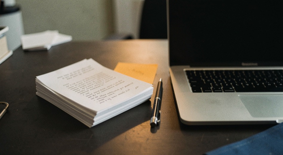 Documentary photograph of a small stack of paper drafts, a mechanical pencil and a compact laptop on a dark desk, no readable words, a single warm pool of light. Distinct supporting card P111 3 composition for this editorial entry.