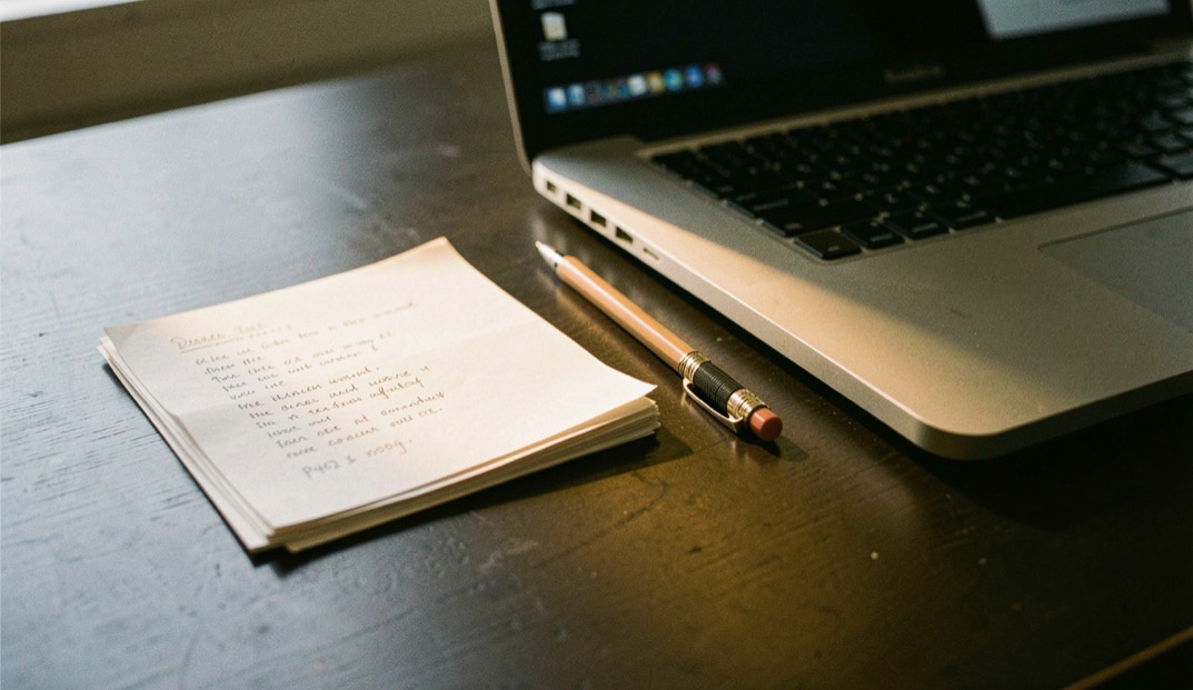 Documentary photograph of a small stack of paper drafts, a mechanical pencil and a compact laptop on a dark desk, no readable words, a single warm pool of light. Distinct supporting card P402 3 composition for this editorial entry.