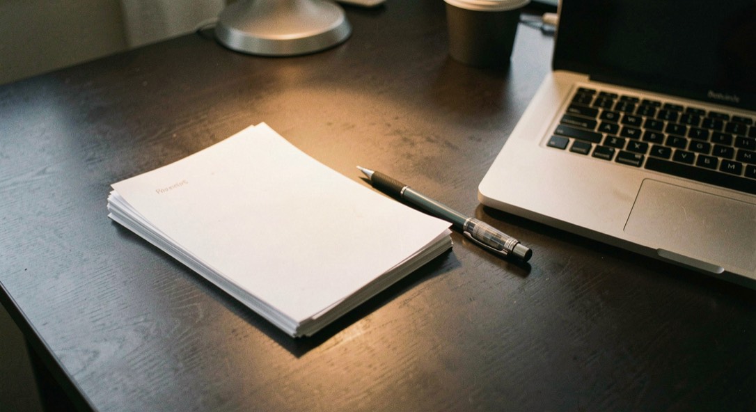 Documentary photograph of a small stack of paper drafts, a mechanical pencil and a compact laptop on a dark desk, no readable words, a single warm pool of light. Distinct supporting card P405 3 composition for this editorial entry.