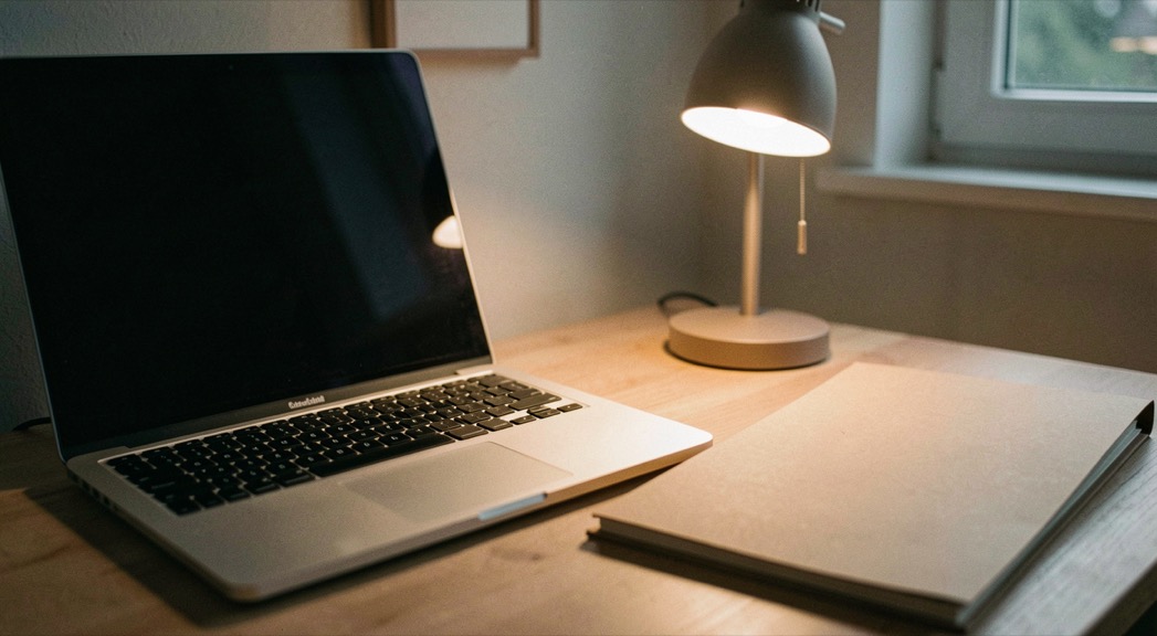 Documentary photograph of a laptop keyboard, a paper folder and a small desk lamp in a calm home workspace, screen turned away, no readable text, evening light. Distinct home journal 8 composition for this editorial entry.