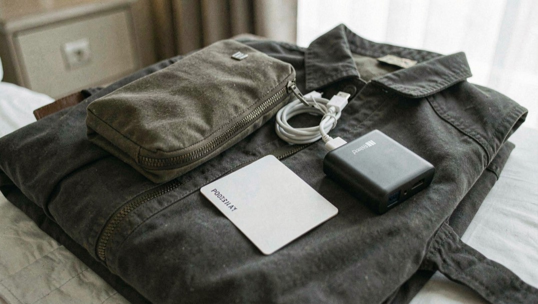 Documentary photograph of a travel pouch, charging cable, passport-sized blank card and compact power bank on a folded jacket, no readable text, hotel daylight. Distinct supporting card P003 3 composition for this editorial entry.