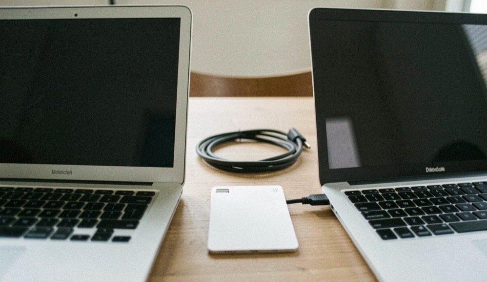 Documentary photograph of two different laptops beside a shared blank data card and a cable, screens turned away, no logos or writing, neutral daylight. Distinct lead composition for this editorial entry.