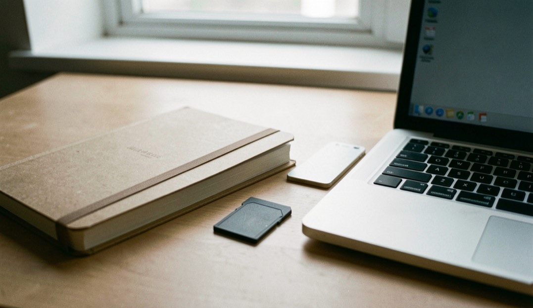 Documentary photograph of a notebook, export folder and blank memory card beside a laptop, no readable screen or writing, quiet window light. Distinct lead composition for this editorial entry.