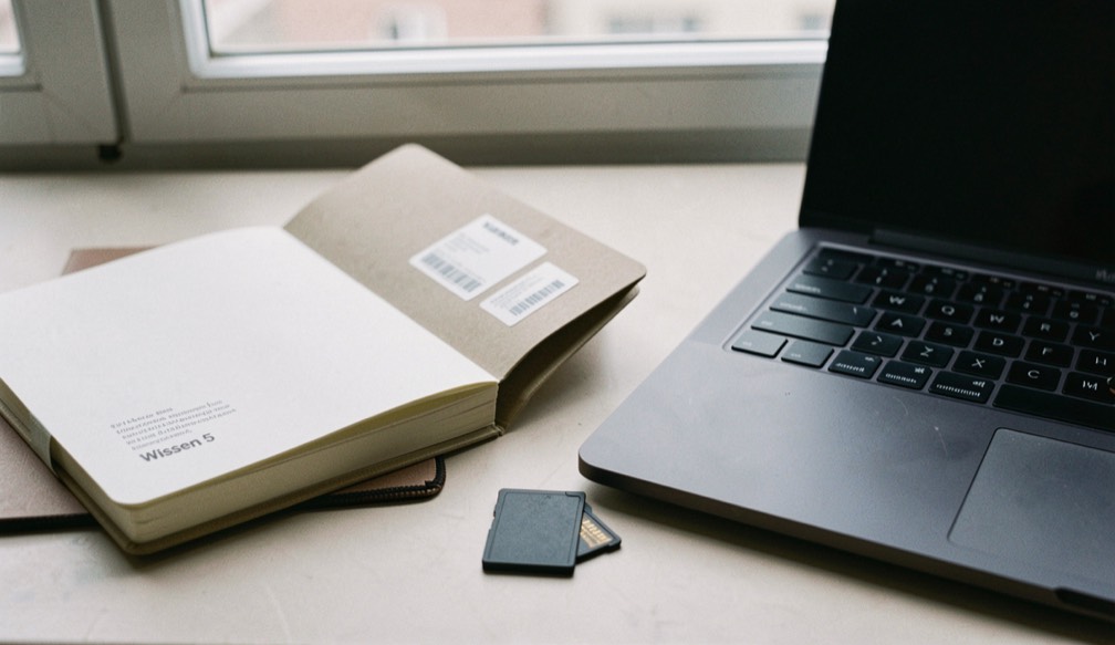 Documentary photograph of a notebook, export folder and blank memory card beside a laptop, no readable screen or writing, quiet window light. Distinct section Wissen 5 composition for this editorial entry.